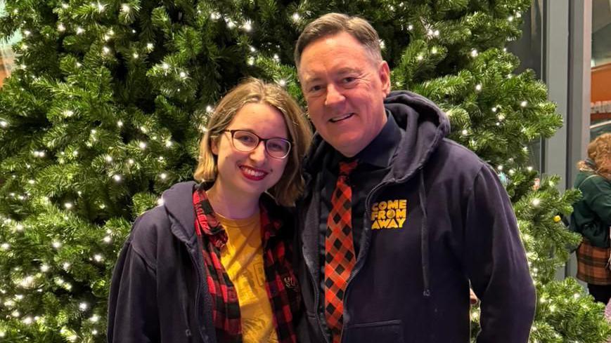 Lydia Greatrix is looking at the camera and smiling. She is stood in front of a green Christmas tree and is wearing a yellow t-shirt, a red checked shirt and blue zip up hoodie. She has a blonde bob and wears glasses. Stood next to her is Kevin Tuerff who is looking at the camera and smiling and is wearing a blue Come From Away zip up hoodie and a red and black checked tie. 