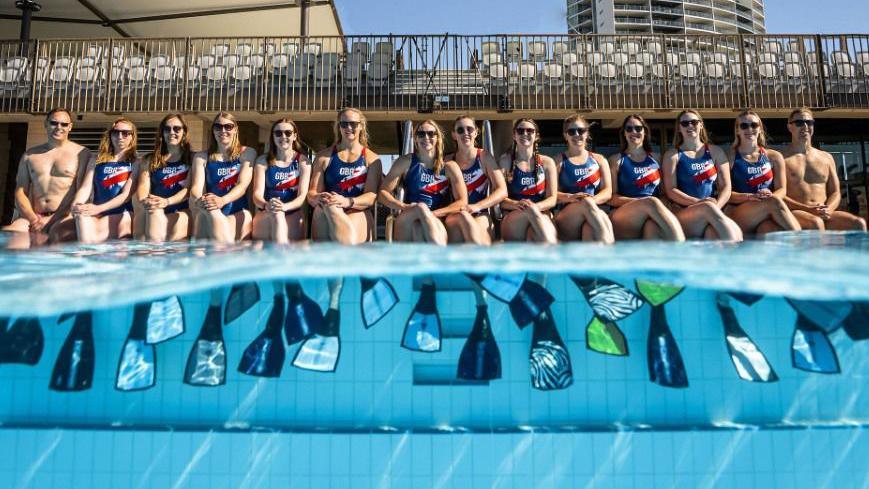 A group of people in red, white and blue swimming suits sat by the edge of an outdoor pool. The group are sat wearing flippers in the water. Half of the shot is above the water, while the other half is underwater.