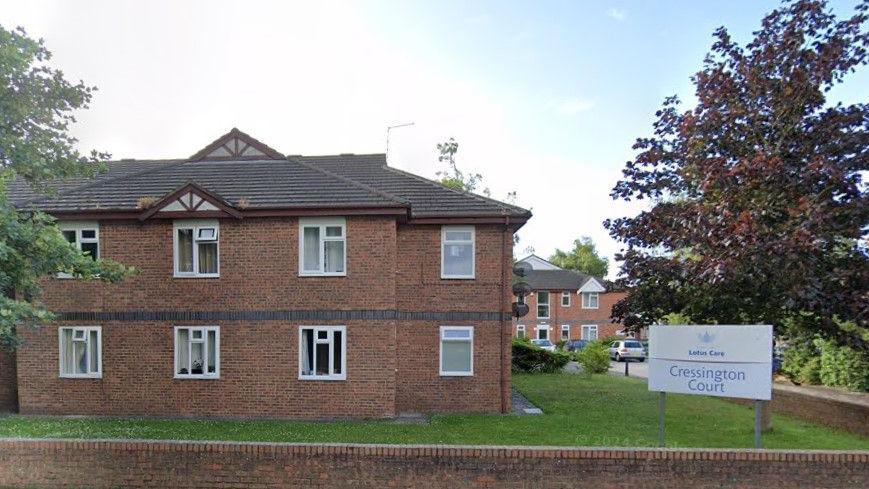 Cressington Court Care Home is a two-storey brick building surrounded by a lawn and trees. Its noticeboard is in the bottom right of the shot.