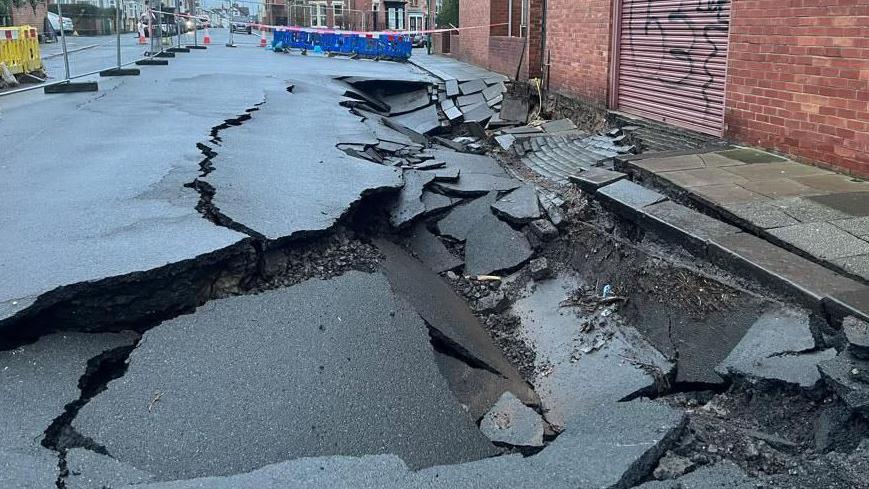 Grey temporary fencing and fluorescent barriers surround the sinkhole, pictured in January. The grey tarmac road is splintered and sunken.