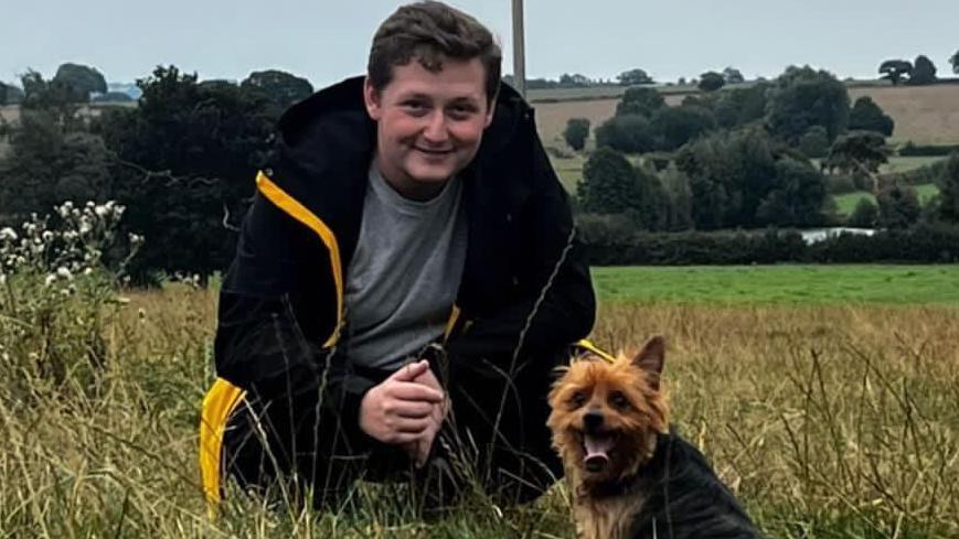 Harvey Denton wearing black anorak with yellow linings in a field with Yorkshire Terrier Pip.