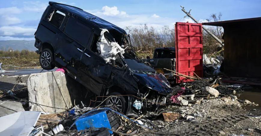 A black SUV is hanging off the side of the road, with its front smashed in and windscreen missing. It is surrounded by other detritus, which includes a red door, but the building around it almost completely disappeared. A fallen telegraph pole is in the background and some sea can be seen beyond the other side of the road. 