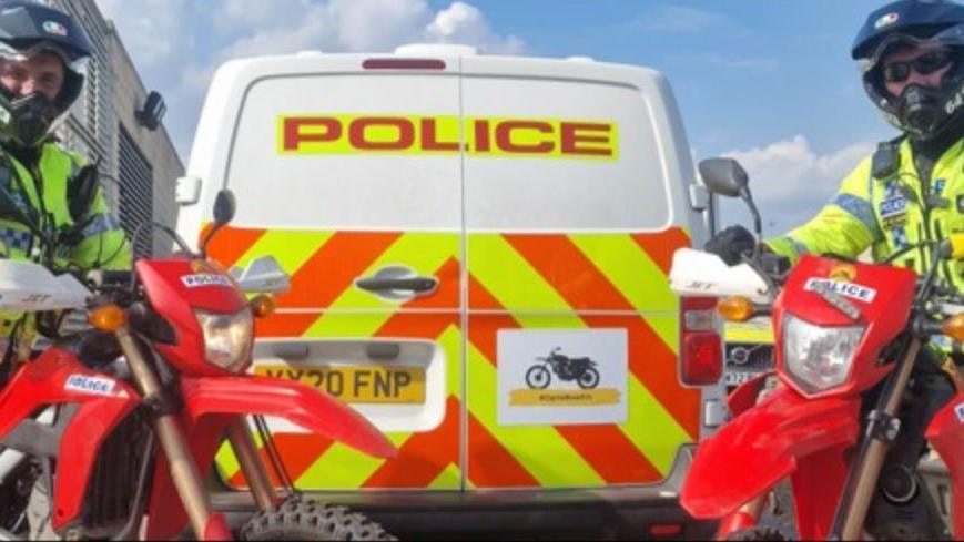 The back of a police van marked with yellow and orange stripes. To the right is a red police bike with an officer sat on it wearing a black helmet, black sunglasses and a yellow hi-visibility jacket. The sky is blue with some clouds.