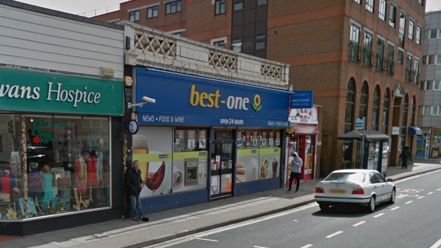 A shop on a high street with a blue sign which reads "Best One". There is a car parked on the road outside.