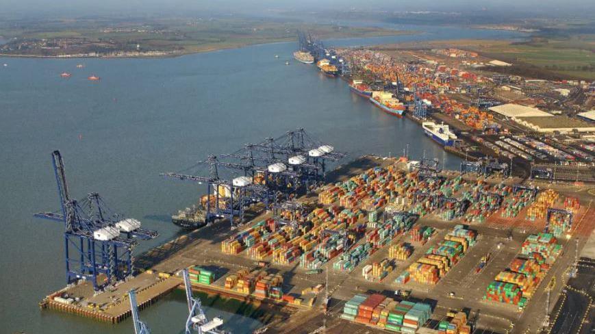 An aerial view of the Port of Felixstowe, showing cranes, containers stacked on the quays and half a dozen ships docked. In the background are is the village of Shotley Gate and the River Orwell.