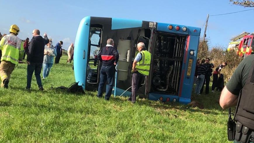 Blue bus overturned on a field, there are crowds of people and the bus windows have been smashed through on impact. Two men are standing in front of the bus.