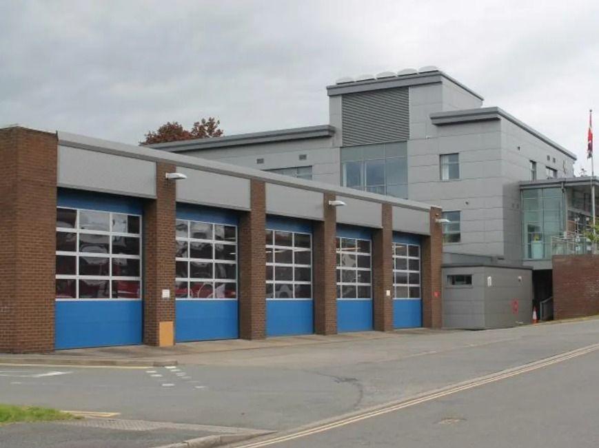 A fire station pictured from the road outside. A high appliance bay can be seen, with four large doors that are blue in between brick pillars. A large grey building is on the right.