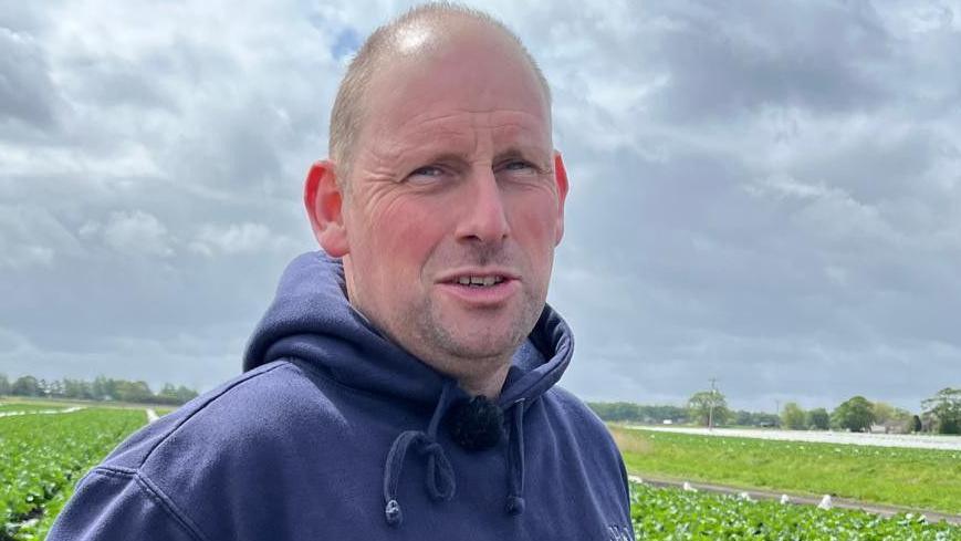 Jon, who is wearing a navy hoodie, is standing in a field of crops with his hands on his waist.