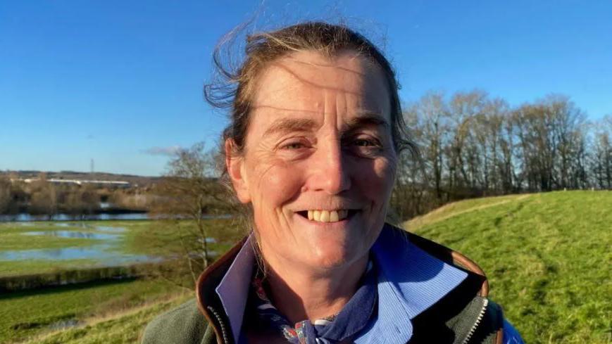 Head and shoulders shot of Amy Wheelton on her farm in South Derbyshire, She is wearing a green coat and is stood in some green fields.