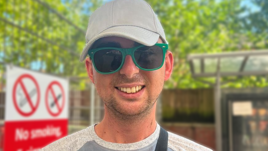 Luke Champeny is facing the camera, smiling. He is wearing dark sunglasses with a green frame, a light grey baseball cap, and a grey top.