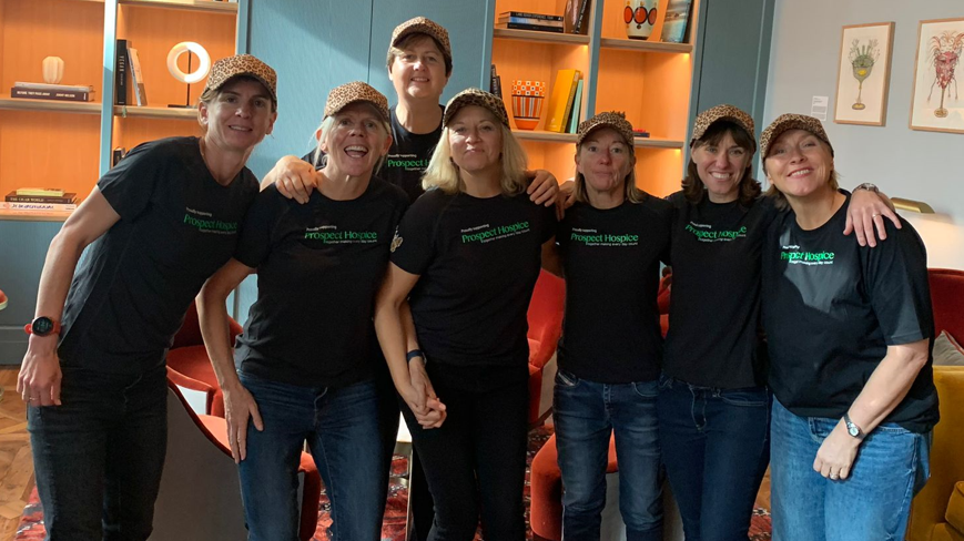 The five women and two of their support team, in a line-up, smiling to camera, wearing black t-shirts with the Prospect Hospice logo on the front and leopard print baseball caps, standing in a large hotel-type room 