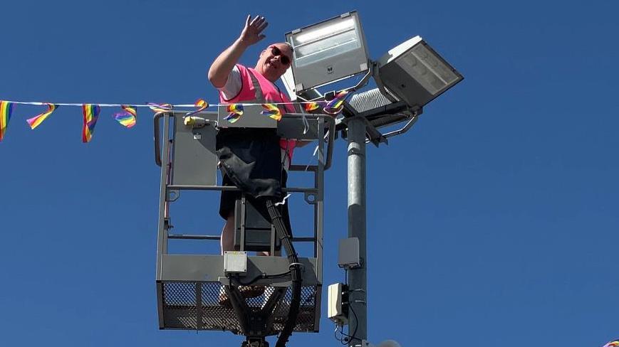 A man in pink high-vis waves whilst standing in a cherry picker tying bunting to a lamppost