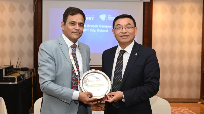 Two men holding a silver plate. On the left a man in a grey suit with a red tie, on the right a man in black suit, black tie and glasses. The photo is taken in front of a presentation. 