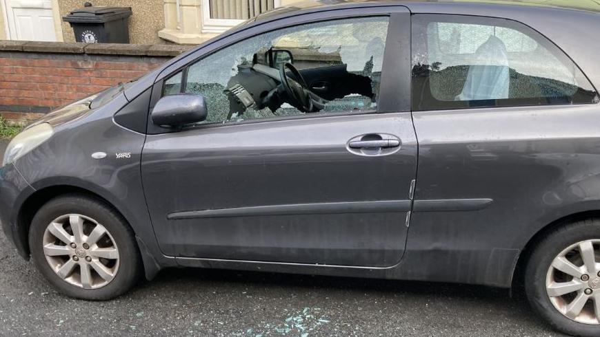 A dark grey car parked on a street in daylight with its window smashed. Broken glass can be seen on the floor. 