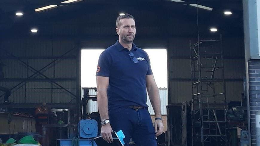 Carl Honeywood wearing a navy blue polo shirt and denim jeans while standing in front of an open industrial unit. He is holding a black notebook and pen and looking into the distance.