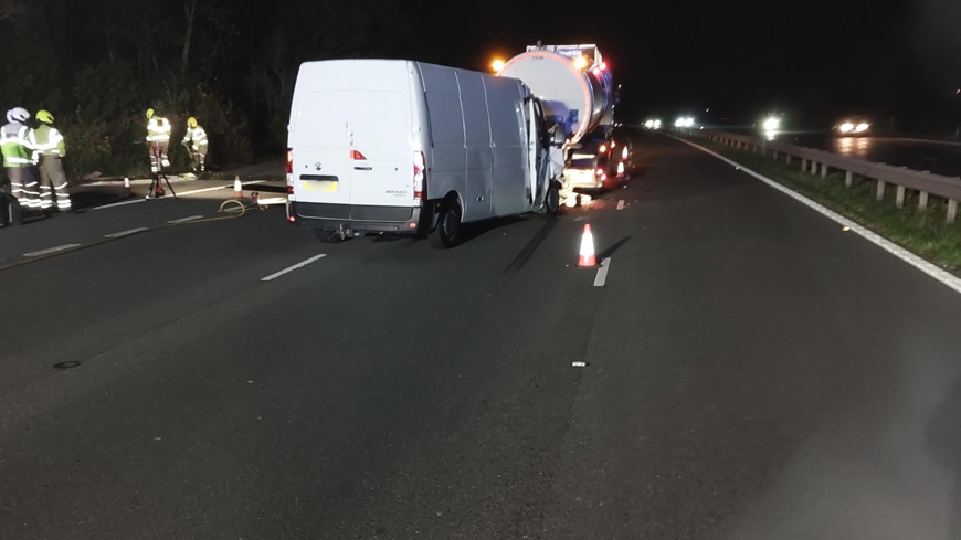 White van behind a tanker in lane two of a three lane motorway following a crash. It is night. To the left several firefighters are putting up large lights.