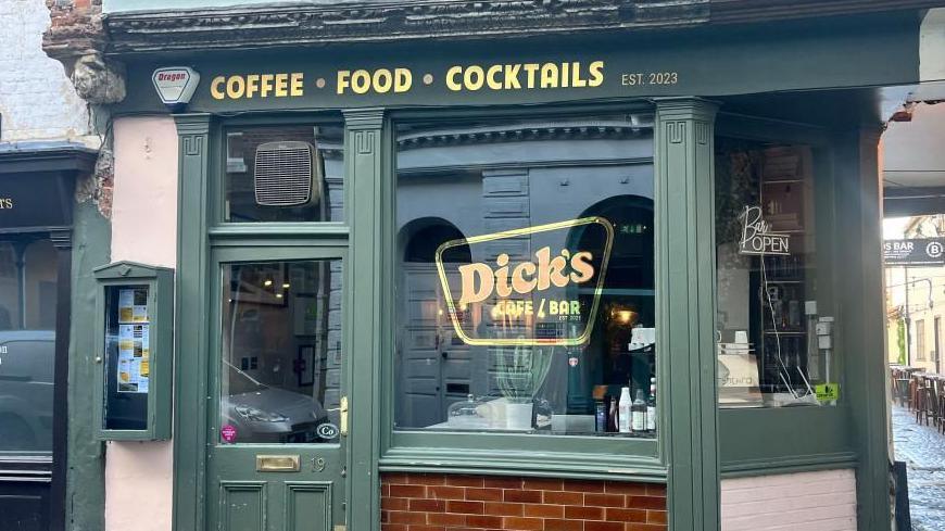 The front of a café. There's a door on the left, and a big window on the right. The area is painted green, and the window has a sign for Dick's Café and Bar. Above the door and window, there is a sign with gold lettering. 