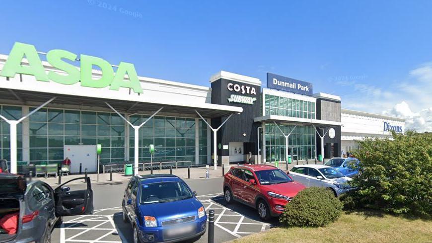 A general view of Dunmail Park from its car park. There is an Asda store to the left, with floor-to-ceiling paned windows. To the right is the entrance to Dixons furniture store, which is all white with a blue sign. Cars are parked in the car park in the foreground.