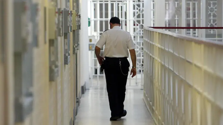 a prison officer in uniform walking away from the camera along a prison wing of cells