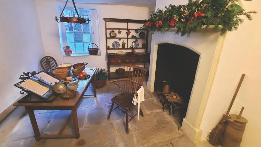 A room inside Charles Wesley's home. There is a kitchen table with some chairs placed next to it. There are some bowls, utensils and a large book on top of the table. There is a fireplace in the room with some wreathes laid on the mantlepiece. In the corner there is a cabinet which is decorated with plates and mugs.