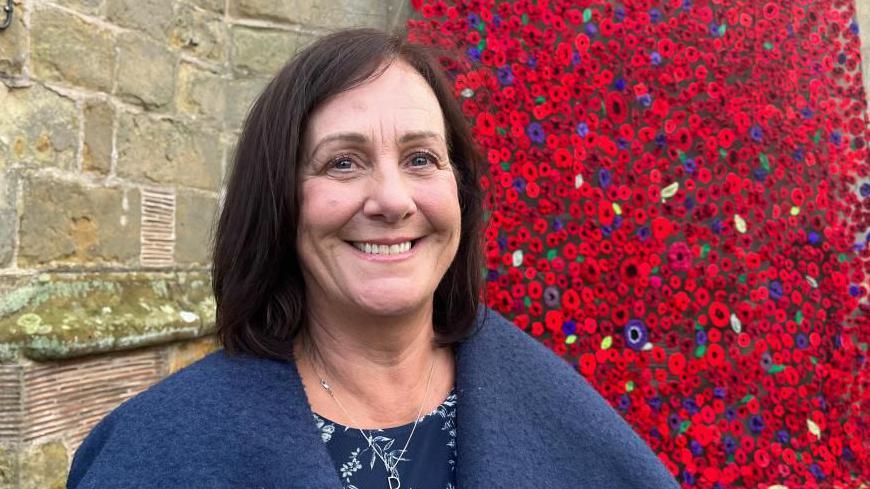 Donna pictured looking into the camera and smiling. She's wearing a navy blue dress with a white floral pattern, and a lighter blue wool coat. She's stood in front of the bottom of the cascade - a wall of knitted poppies in red and purple. 