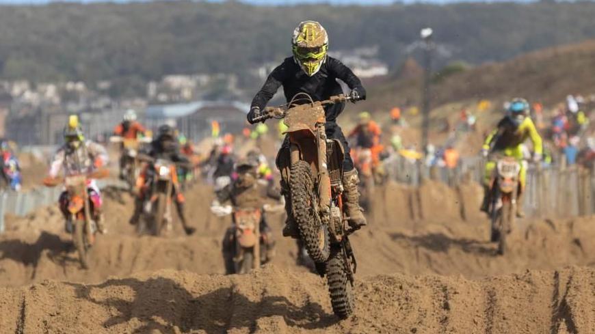 A dirt bike rider wearing black clothing and a yellow helmet performs a jump during the Weston Beach Race. Dozens of other riders can be seen following behind, with Weston Super Mare in the far background. All riders are clearly splattered with sand.