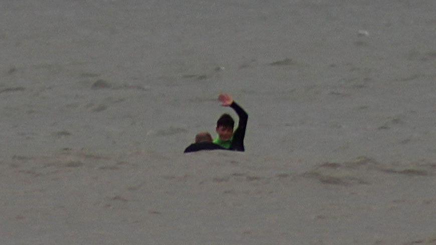 Oscar is in a grey sea signalling with his arm over his head and the head and shoulders of a man is visible in the water next to him.