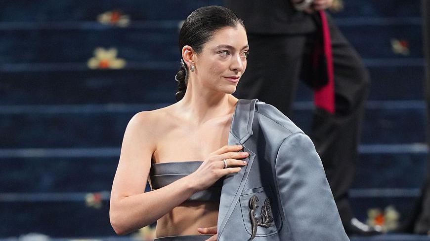 Lorde at the Met Gala in New York. The singer wears a grey-blue silk blazer over a backless bandeau top and tight skirt. Her dark hair is pulled back in a plait and she wears a simple make-up.