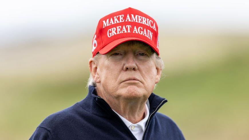 Head and shoulders of Donald Trump, who is wearing a white polo shirt, a navy blue fleece and a red cap which says "Make America great again"
