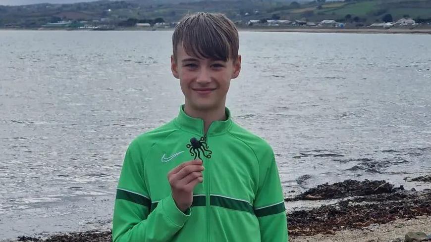 A young boy, wearing a green Nike zip up hoodie, holding a Lego octopus piece. Behind him is the sea. 