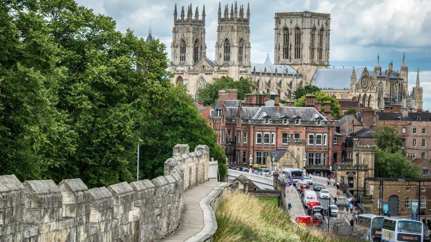 York Minster in the distance with buildings and a road in front of it. City walls to the left of the image with trees behind the walls.