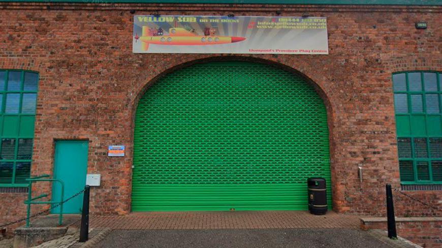 A brick building with door covered with a green metal shutter. A sign above the door reads "yellow sub on the docks" and has a picture of a yellow submarine with cartoon characters sitting on it