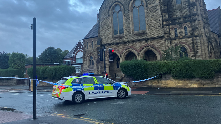 The scene in Moss Side, showing a police car and police cordon in front of a large church building
