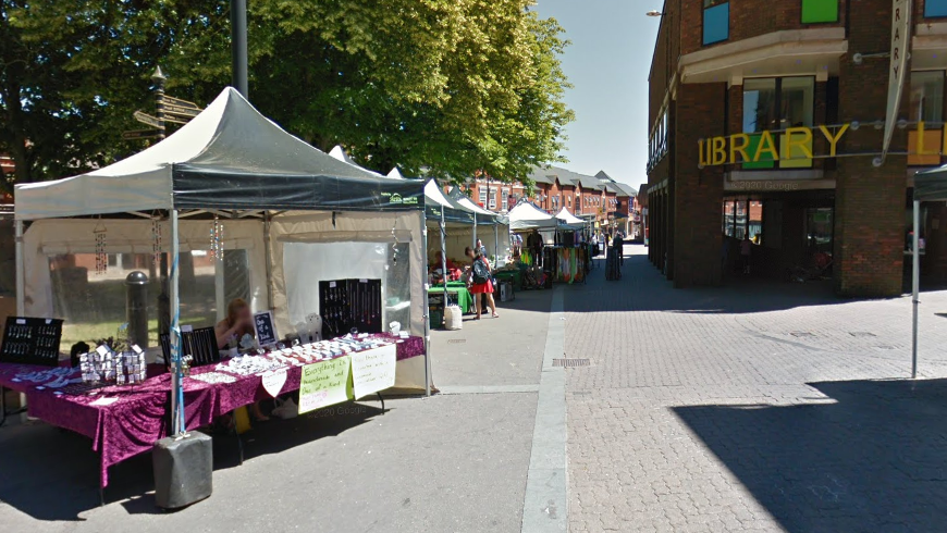 A street with several white market stalls on the left hand side selling products. On the right is a large building with a yellow sign that reads "library". There are green trees behind the market stalls.