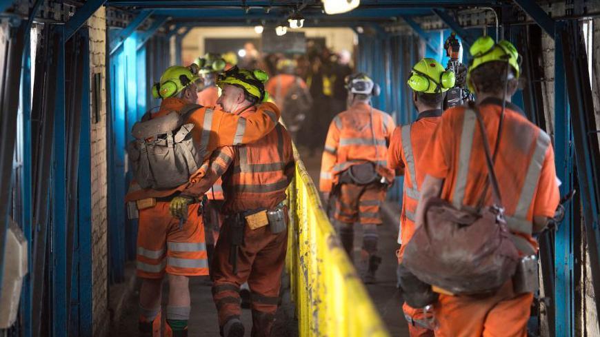 Coal miners wearing orange overalls hardhats and protective equipment hug and engages with each other as they walk through a corridor after finishing their final shift before closure at the Kellingley Colliery in Yorkshire.