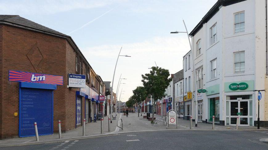 Dalton Road in Barrow is a traditional High Street. A B&M store is on one side in a brown brick building and a Specsavers is on the other in an older white building. The street, which has trees in the middle, is empty bar a couple of people.