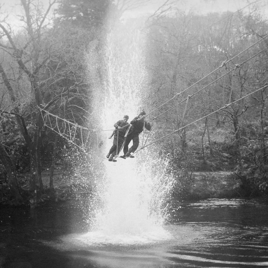 Two commandos carrying rifles cross a rope bridge over water at Achnacarry. A plume of rises up from a small explosion in the water. Trees fringe the water's edge.