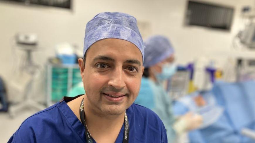 Mr Indy Sian wearing a blue scrub hat and blue scrubs. He is smiling to the camera and behind him is an operating theatre. 
