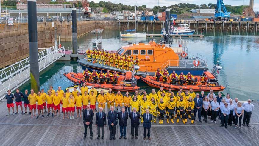 Crew and support staff from the Jersey lifeboat service. The are standing in rows on a pontoon, with several small and large lifeboats behind.