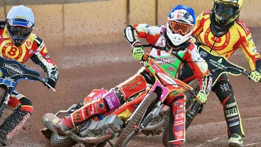 Three speedway riders battle it out on the track. They are wearing multi-coloured race suits 