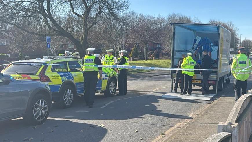 A police cordon on a roadside with a group of officers in uniform talking next to a patrol car. 