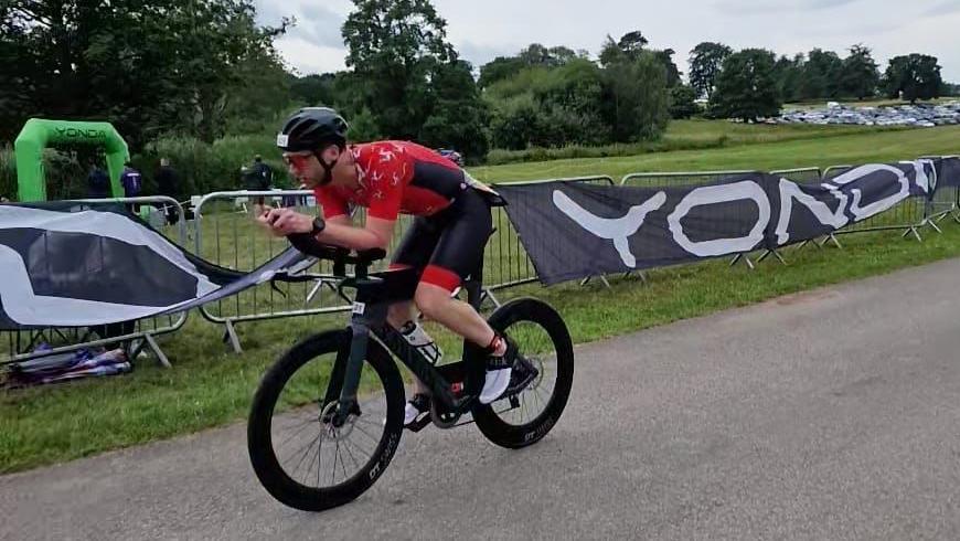 Matt Looker rides a bike on a grey tarmac track by some grass. He wears a red and black shorts and t-shirt and a black helmet.