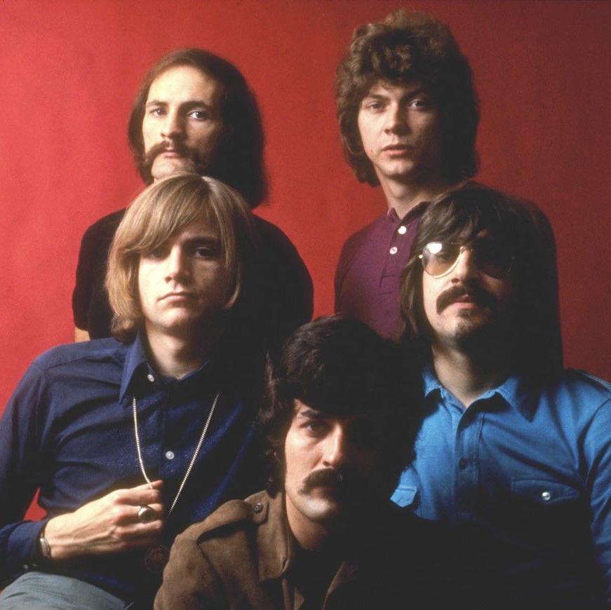 Two men are at the back, with two in the middle and one at the bottom of the image. Three of them have a moustache. Two of them are wearing blue tops, with the others wearing brown, purple and black.