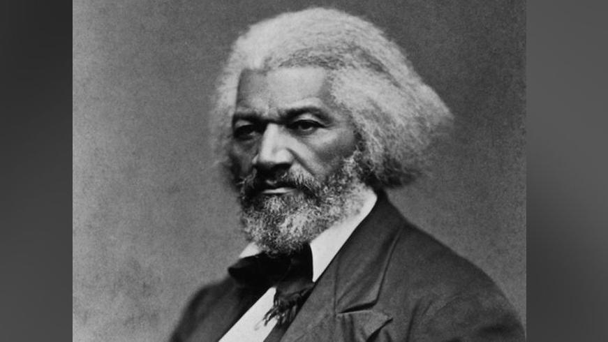 A historic black-and-white picture of Frederick Douglass, wearing a serious expression, with a grey beard, grey hair and a smart suit.