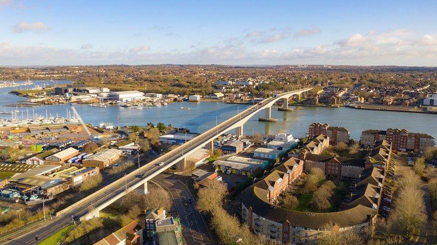 An aerial view of the Itchen Bridge on a sunny day. The two-lane bridge over the River Itchen is in a suburban area with buildings in the foreground. On the far river bank you can see a mix of buildings and trees.