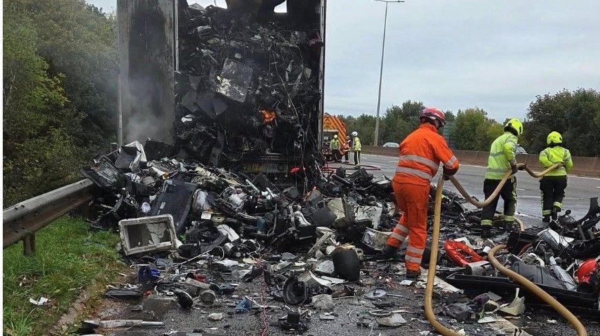 Firefighters battle the flames of a lorry fire on the M5 in Exeter. Two crew members are inspecting breathing apparatus on the road while others work on the fire.