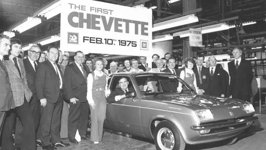 A black and white photo of a Vauxhall chevette car inside the factory. One man sits in the driver's seat and leans out of the window, smiling at the camera. A crowd of about 20 people surround the car in a posed photo, also looking at the camera. A large white sign above the car has the Vauxhall griffin logo and reads: "The first Chevette Feb 10th 1975".