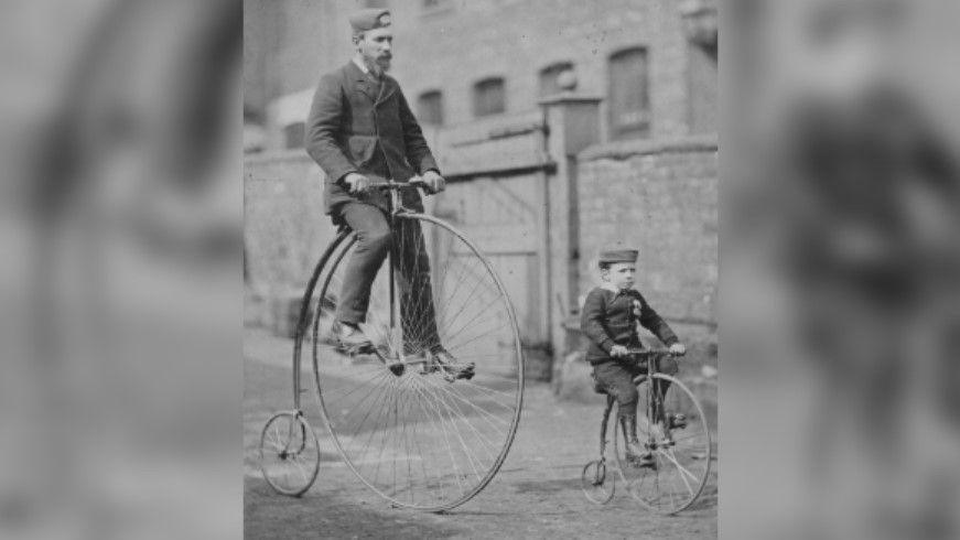 A black and white picture of a man and boy riding matching penny farthing bikes on a cobbled street. They are both dressed in suits and a hat.