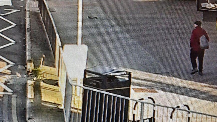 Një imazh i kamerave të sigurisë tregon një burrë që ecën në një trotuar të gjerë pranë një rruge. Ai ka veshur një xhaketë ngjyrë burgundy, një kapelë të errët, pantallona dhe çizme të errëta, dhe mban një çantë mbi njërën shpatull.