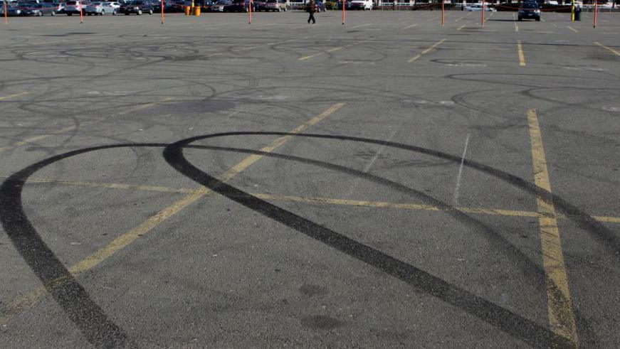 Tire marks in the shape of loops can be seen on the grey tarmac of a car park, with bays marked out by yellow lines.
